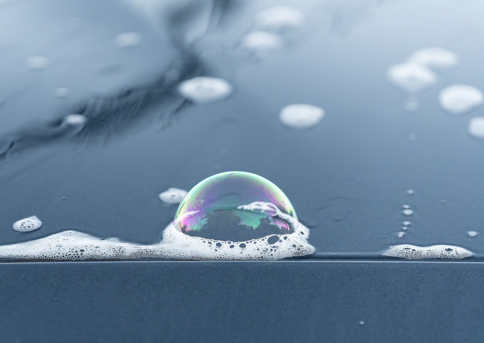 Macro shot of an iridescent soap bubble resting in wash foam on a dark car panel