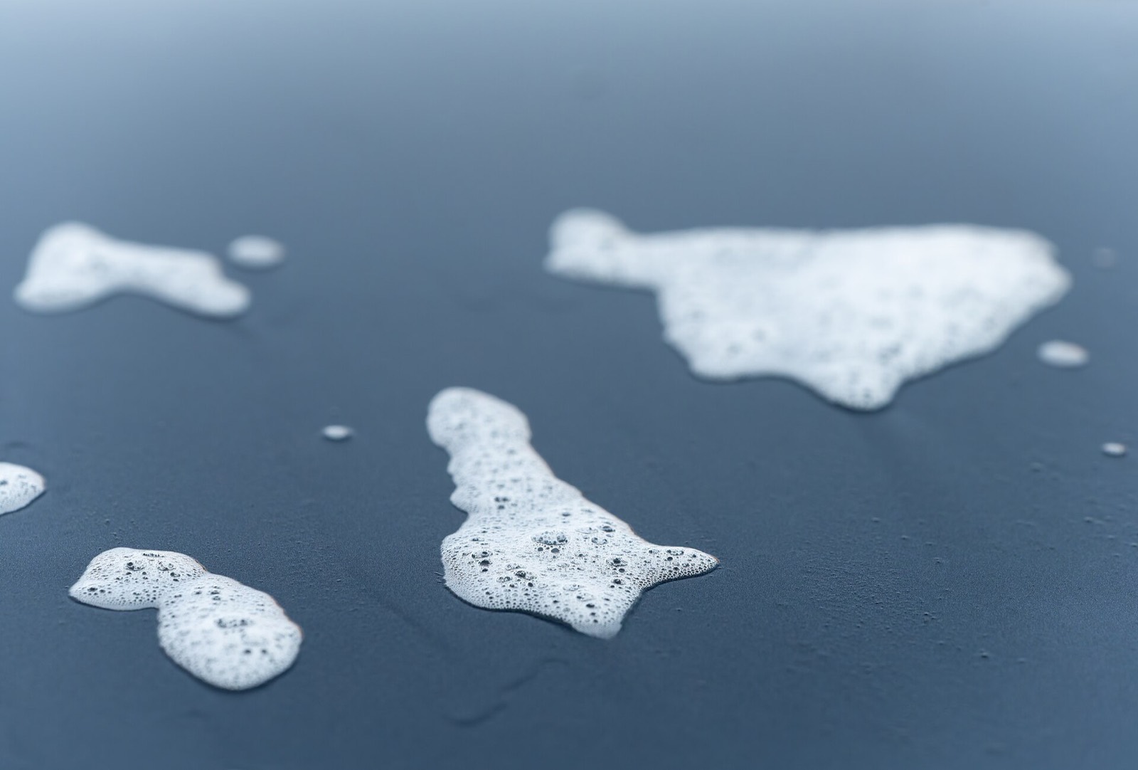 Islands of white wash foam sitting on the wet dark grey paint of a car bonnet